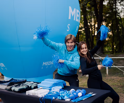 Two young people smiling and holding blue pom-poms, standing behind a table with promotional items in front of a large blue inflatable balloon outdoors.