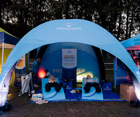 Two people reclining on blue chairs under a blue Medicover Sport tent at an outdoor event with trees in the background.