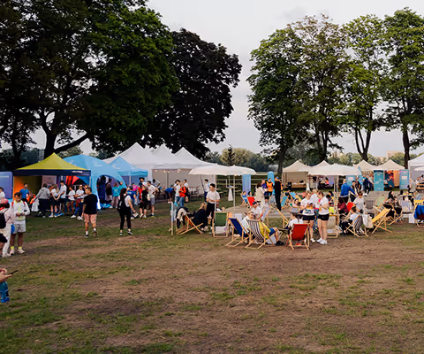 People gathered outdoors at a festival with tents, chairs, and trees in the background.