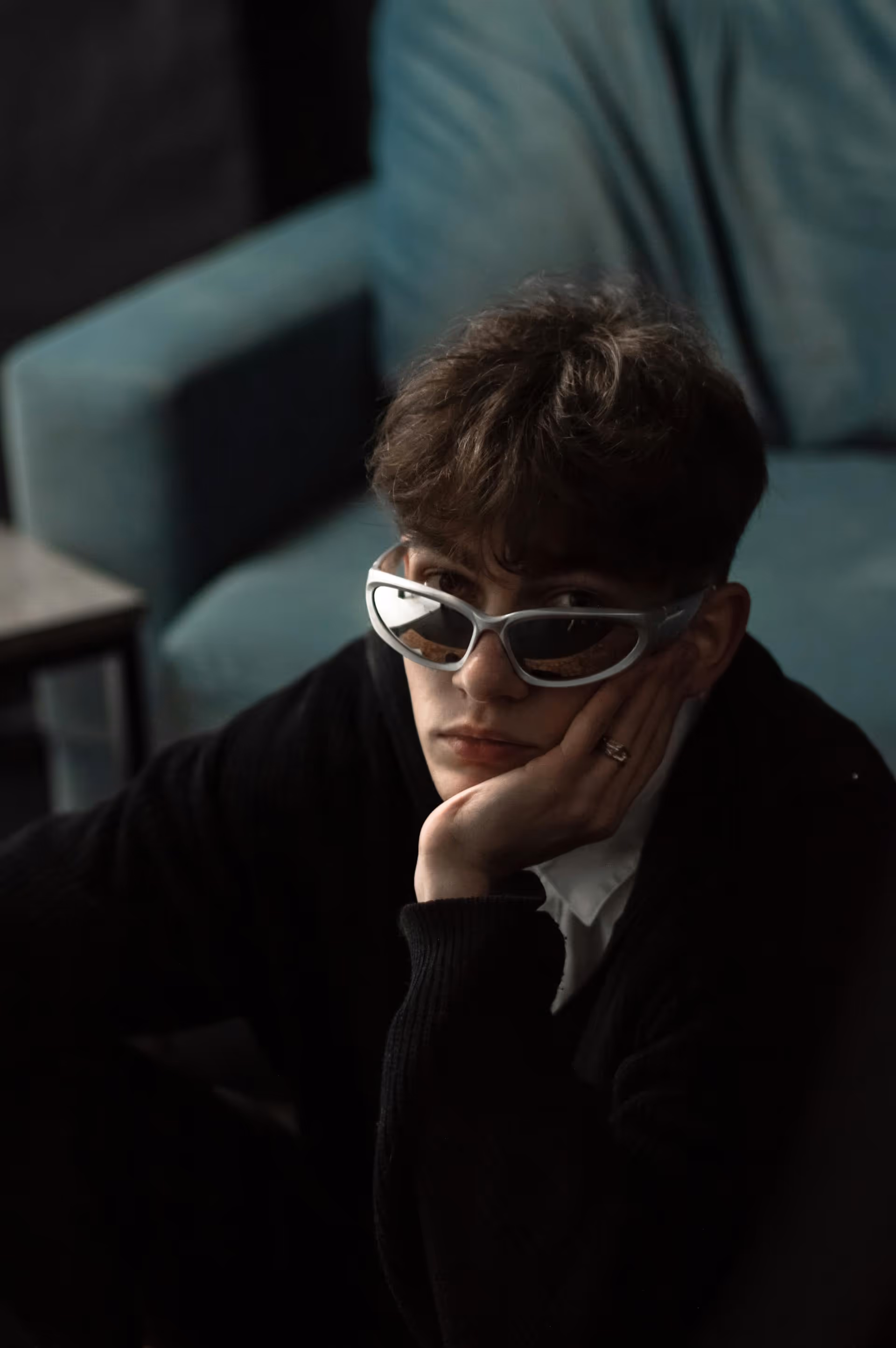 Young person with short curly hair wearing silver-rimmed sunglasses, resting their face on their hand indoors.