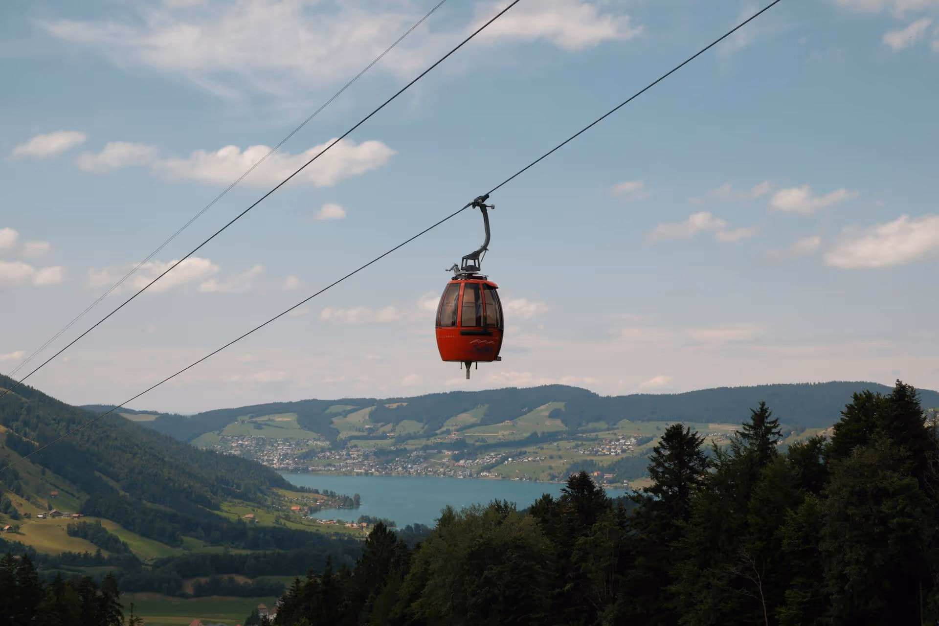 Red cable car suspended on cables above a forested mountain landscape with a lake and distant hills under a partly cloudy sky.