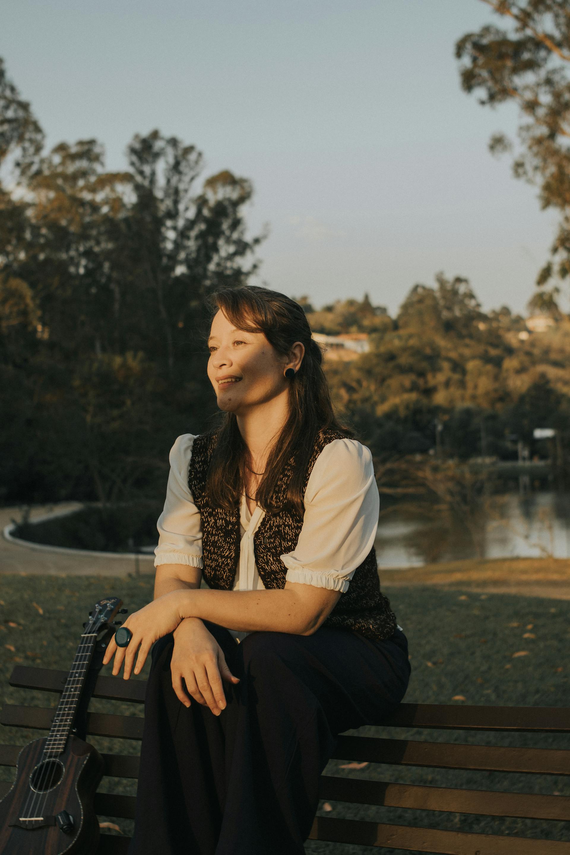 Woman sitting on a bench in a park during golden hour with a guitar resting beside her.