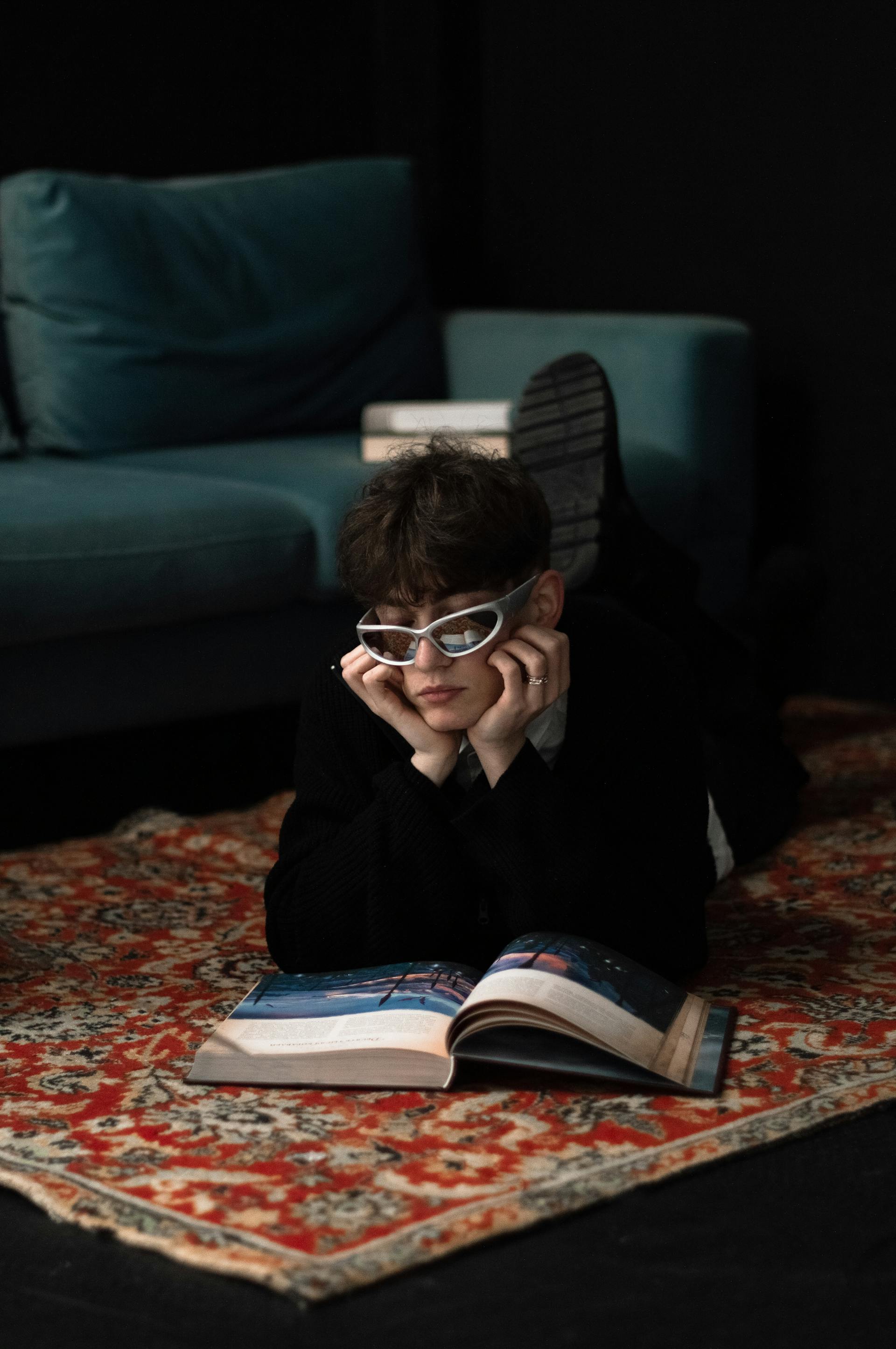 Person lying on a patterned rug reading a large open book with a teal sofa in the background.