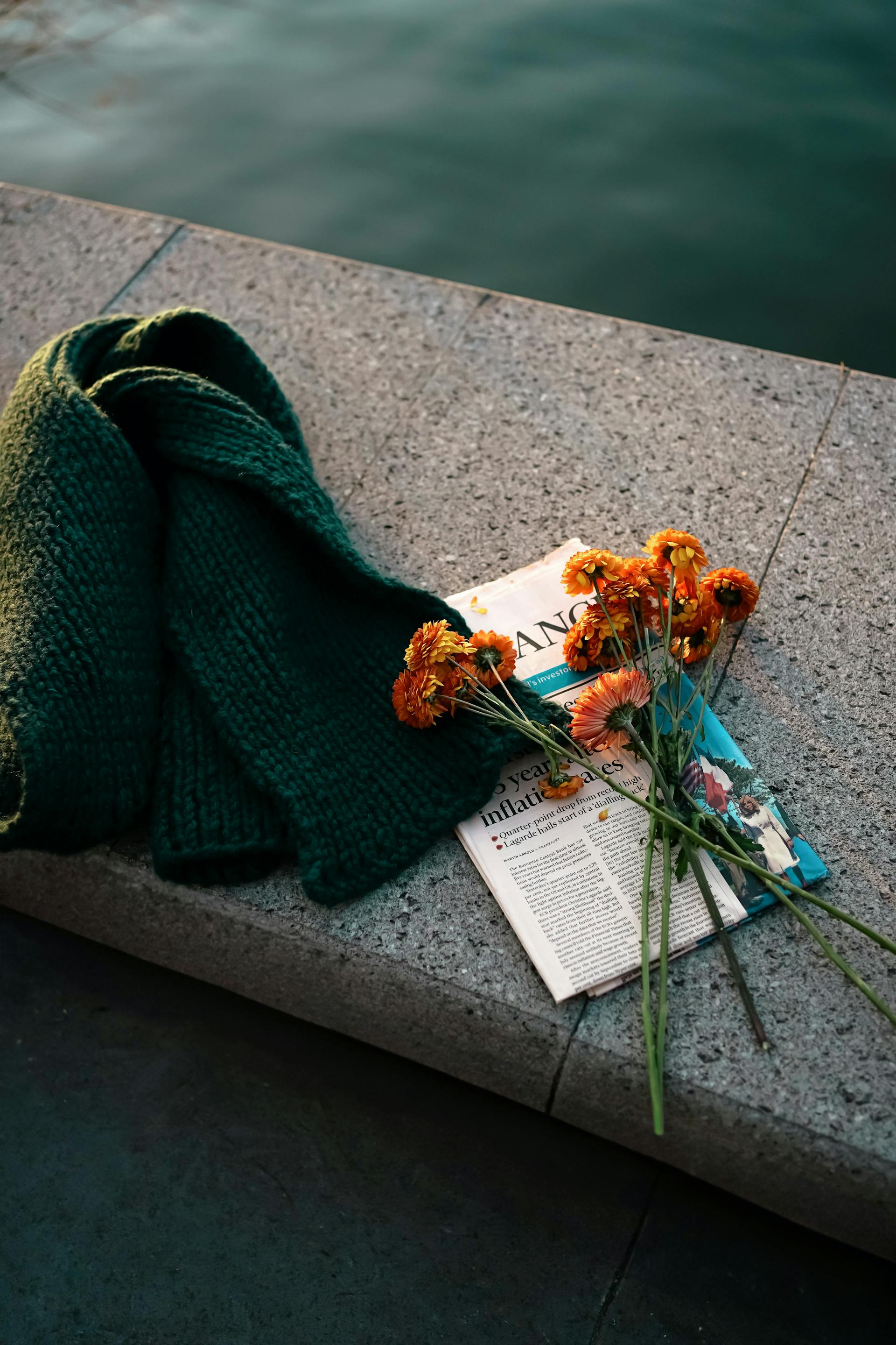 Orange flowers and a dark green knitted scarf resting on a folded newspaper on a stone surface near water.