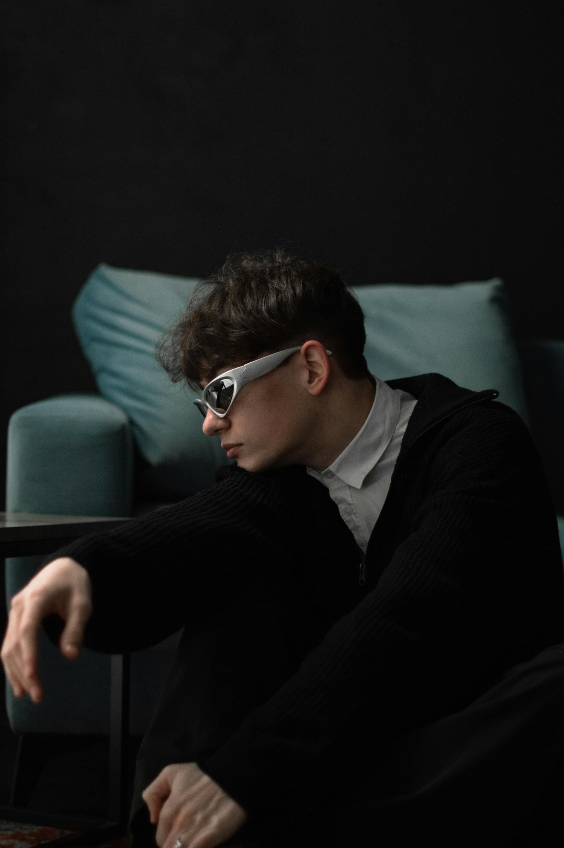 Young person wearing white futuristic sunglasses and a black sweater, sitting indoors with a teal armchair in the background.