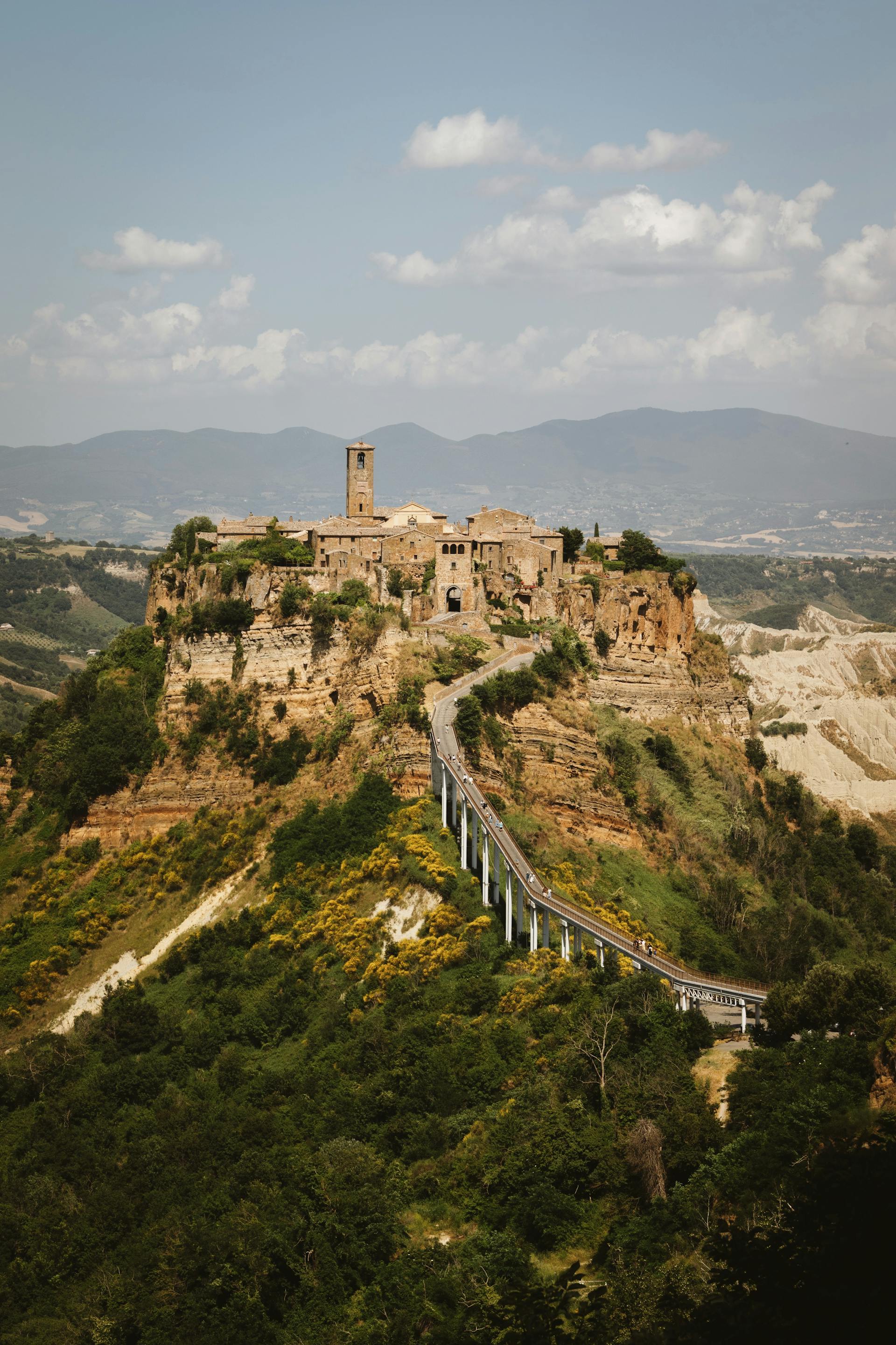 Ancient hilltop village connected by a narrow bridge with mountainous landscape under a partly cloudy sky.