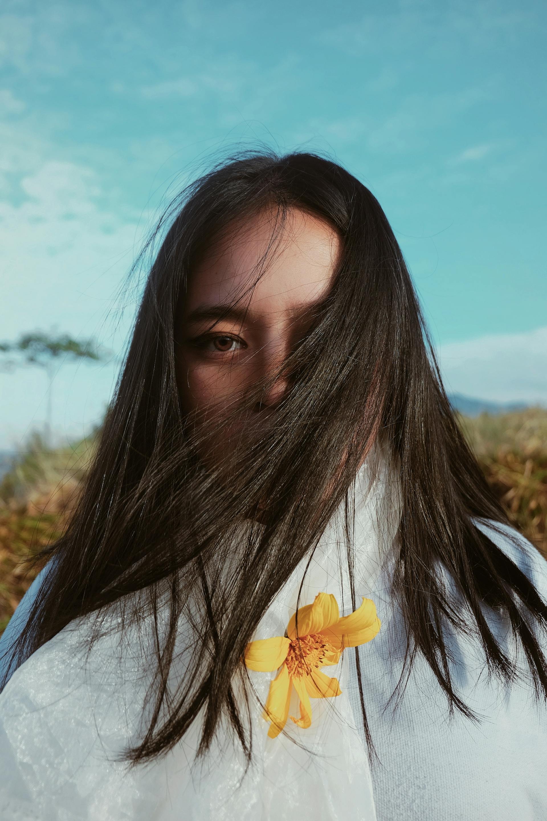 Close-up of a woman with long black hair partially covering her face, holding a yellow flower against a light blue sky.