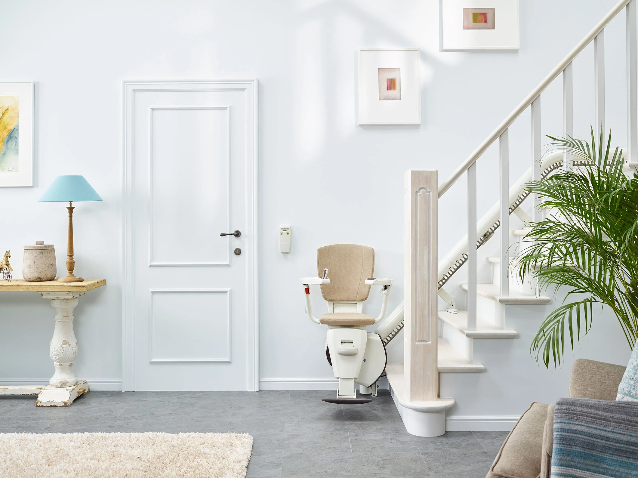 Trapliftstoel geïnstalleerd naast een witte trap in een lichte kamer met meubels en planten.