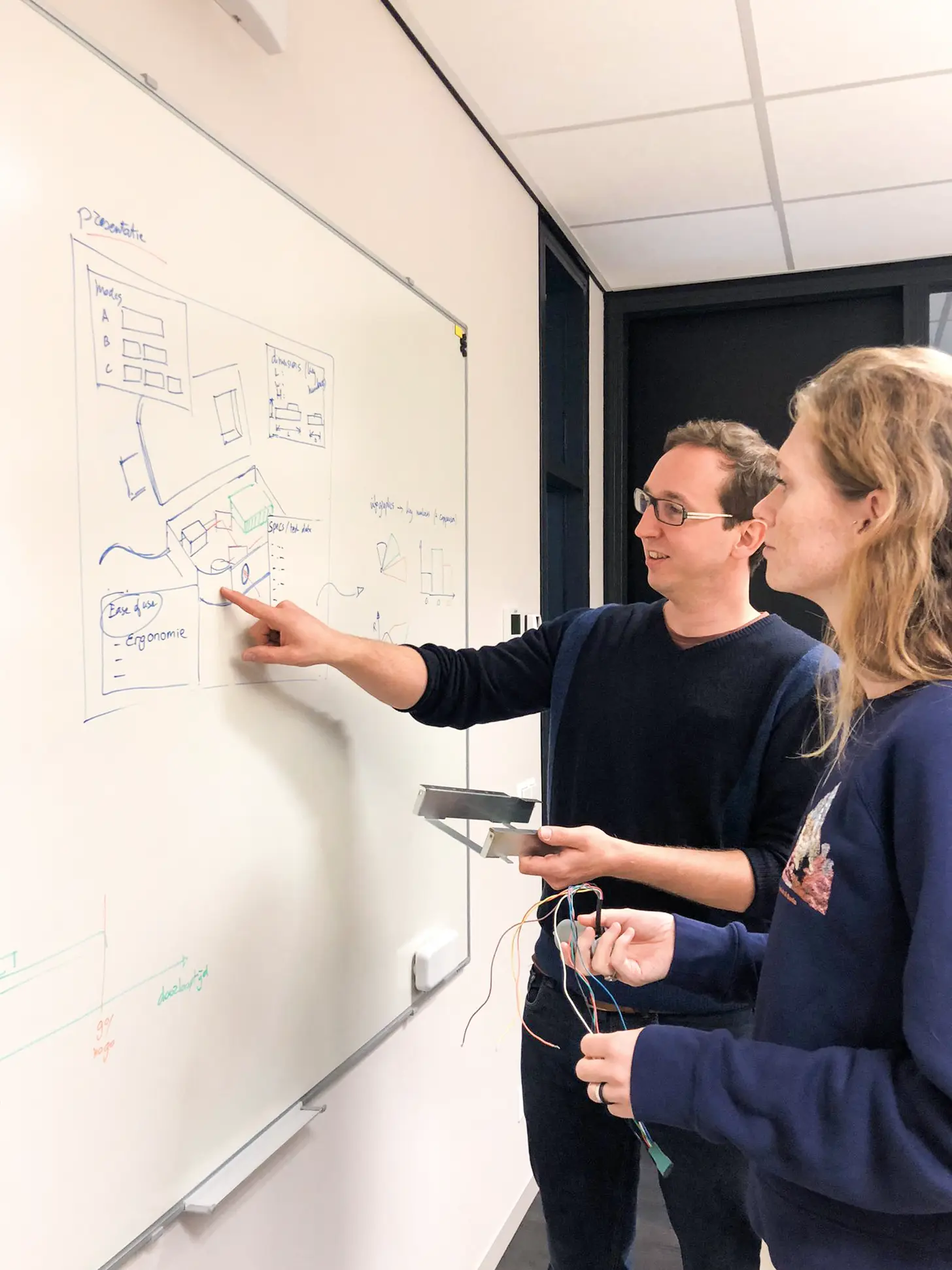 Twee mensen bespreken een diagram op een whiteboard terwijl ze elektronische draden vasthouden.