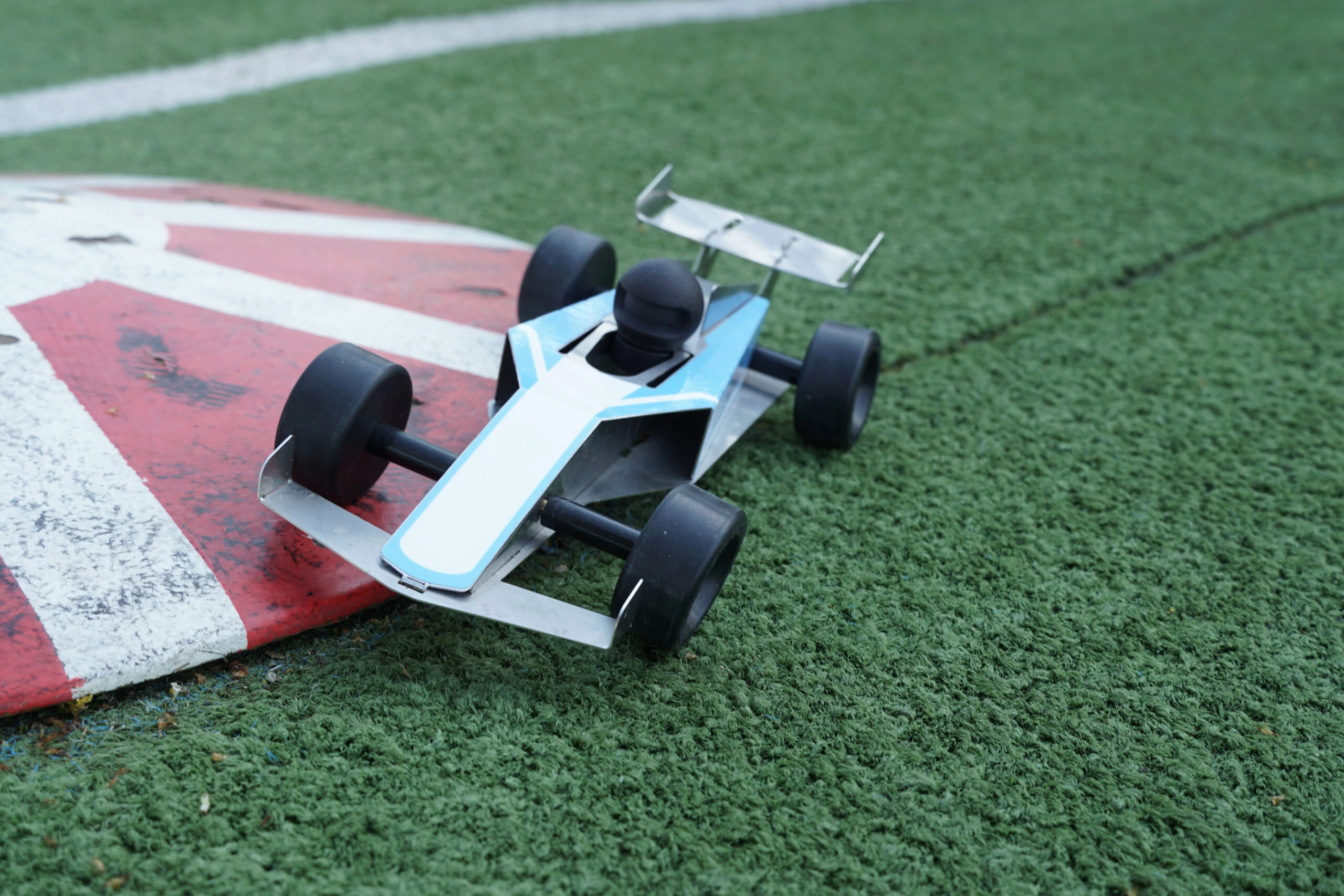 Een kleine modelraceauto op gras naast een rood-witte bochtmarkering op een racecircuit.