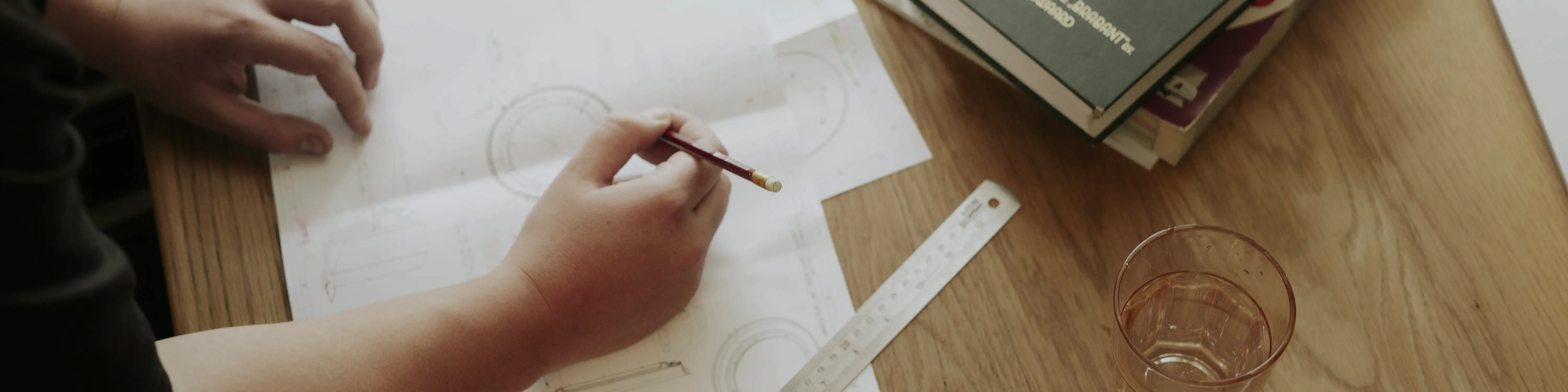 Iemand schrijft met een potlood op technische tekeningen op een houten bureau met liniaal en boeken.