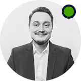 Smiling man with a mustache and short hair, wearing a suit and white shirt, against a light gray circular background.