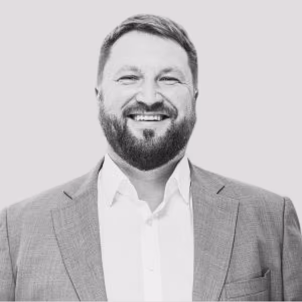 Smiling man with a beard wearing a suit jacket and white shirt against a plain background.
