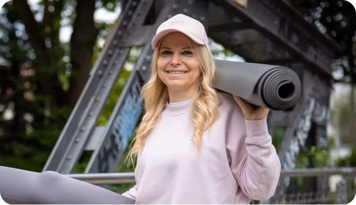 Smiling woman in pink sweatshirt and cap holding a rolled yoga mat on her shoulder outdoors.