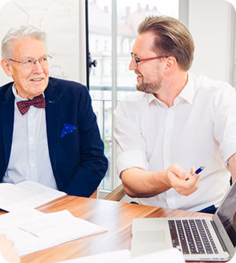 Two men in a business meeting smiling and discussing documents with a laptop on the table.