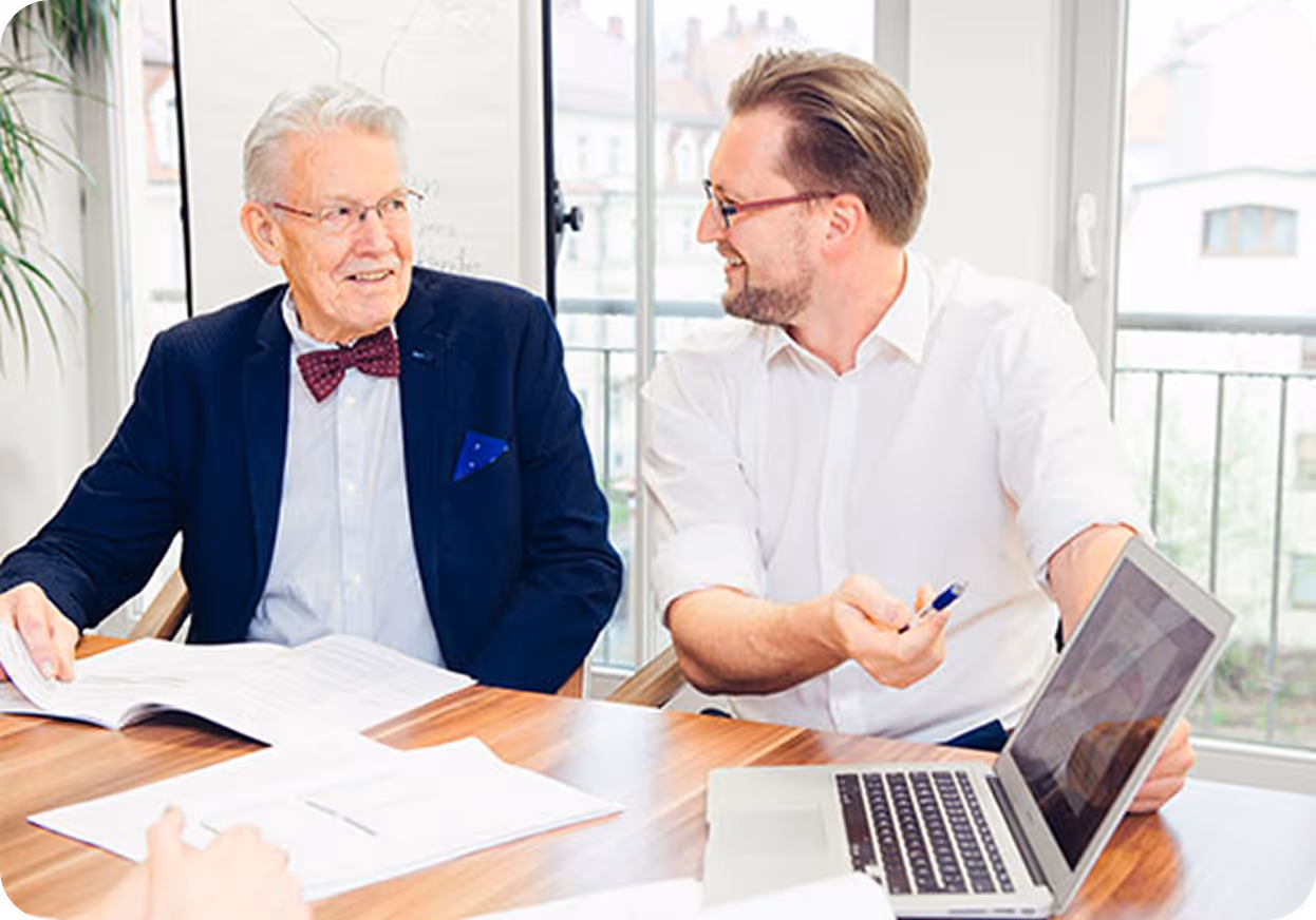 Two men, one older wearing a bow tie and glasses, and one younger with glasses, smiling and discussing documents at a table with a laptop.