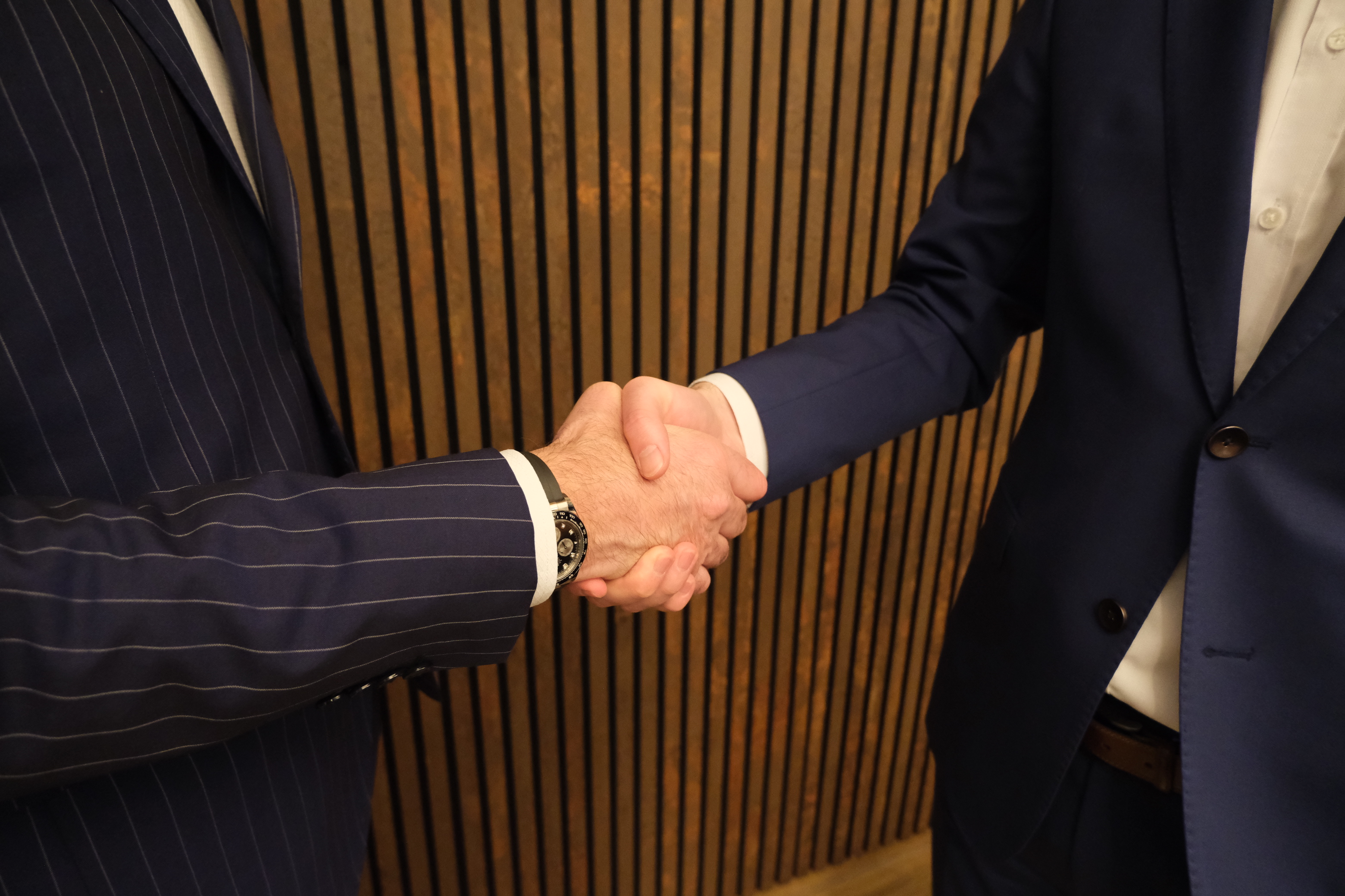Zwei Männer in Anzug geben sich vor einer Holzwand die Hand als Zeichen der geschäftlichen Partnerschaft und Vertrauen.