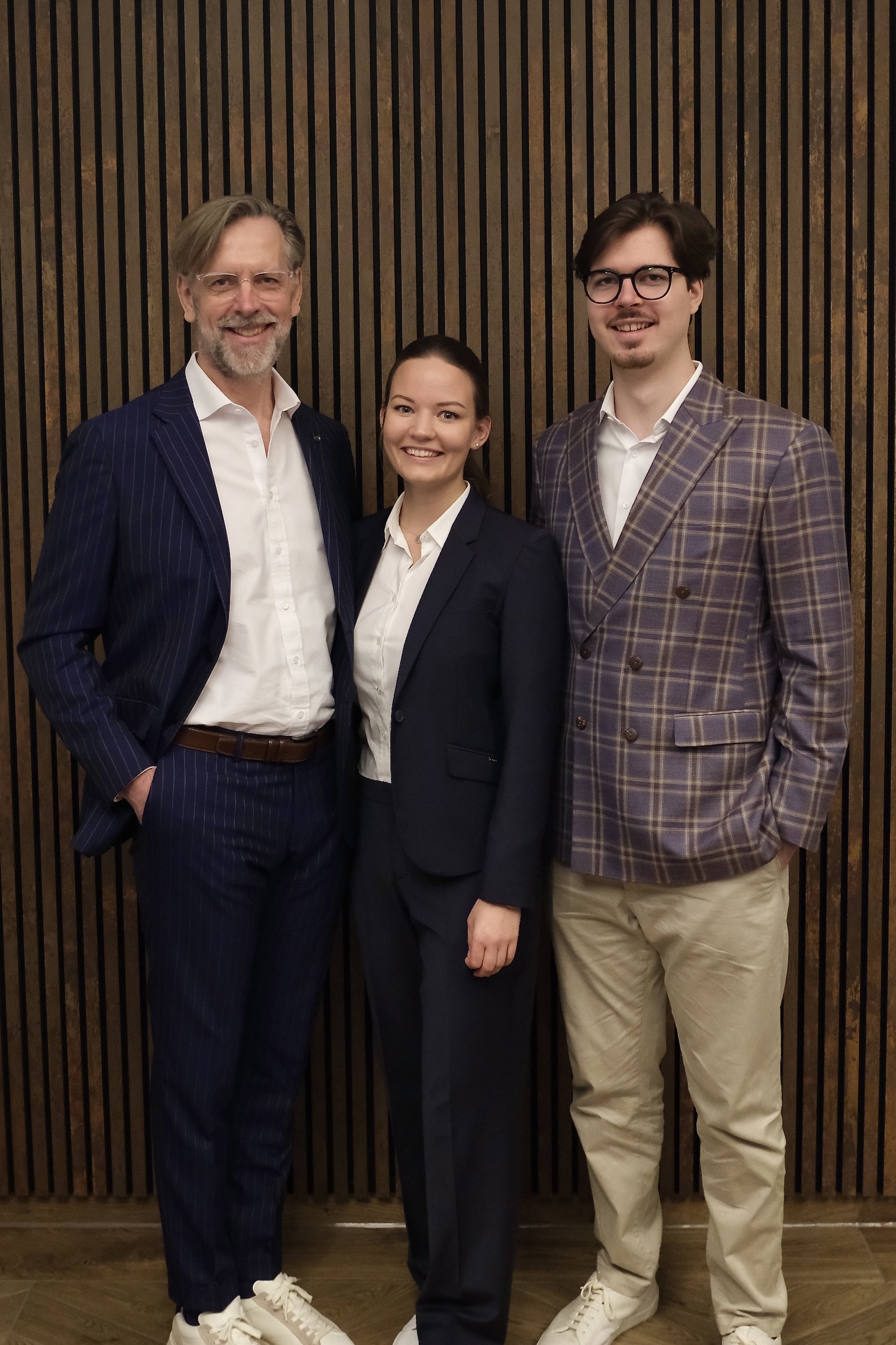 Three people standing in front of a vertical wooden slat wall wearing business casual attire, smiling at the camera; the man on the left, Krzysztof Bialon, ist the Founder & CEO, the woman in the center is Carina Fischinger (Co-Founder, Health & Innovation), and the man on the right is Adrian Bialon (Co-Founder & Managing Director).