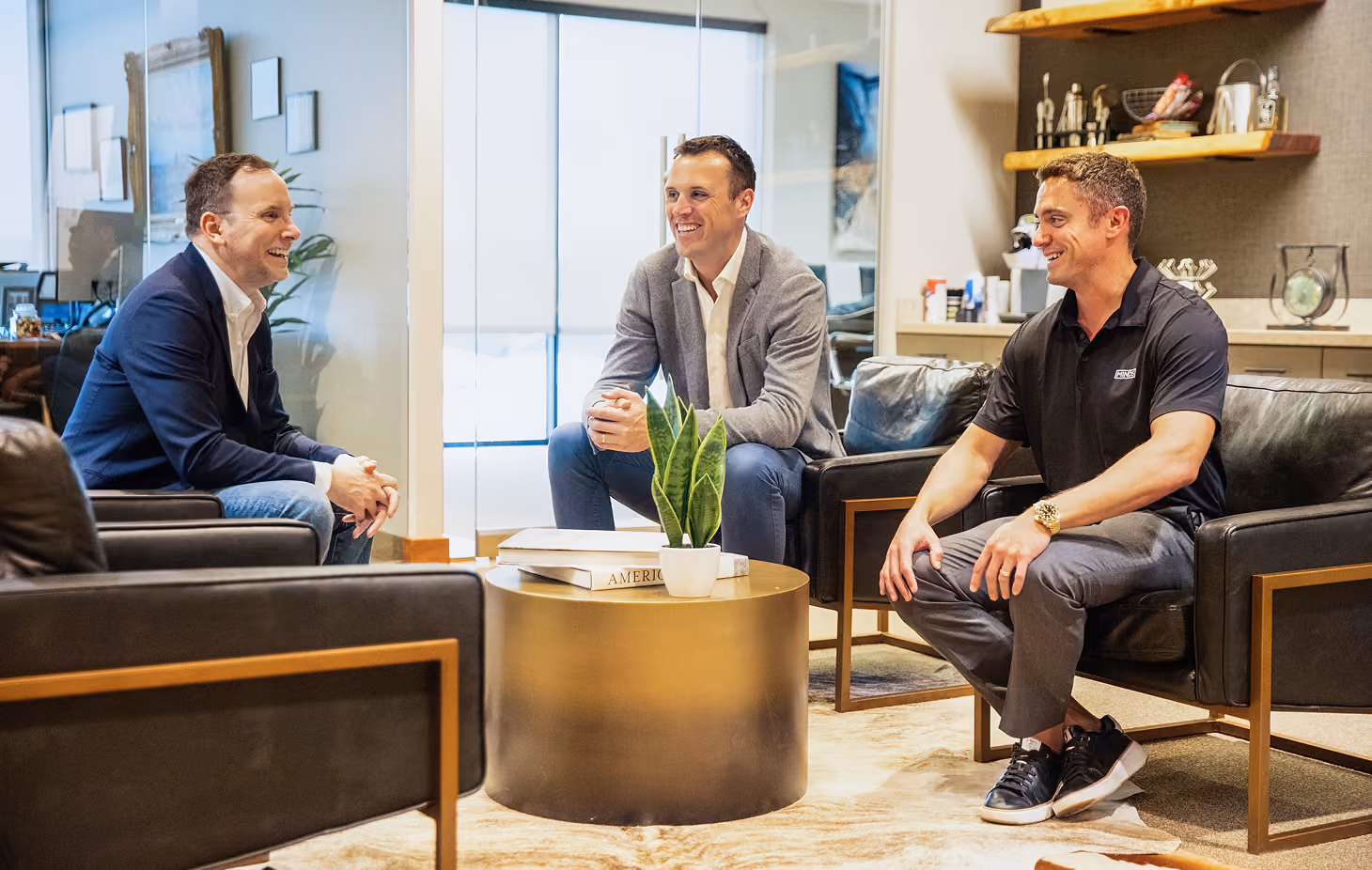 Three men sitting and smiling in a modern office lounge with leather chairs and a round gold coffee table holding a small potted plant and books.