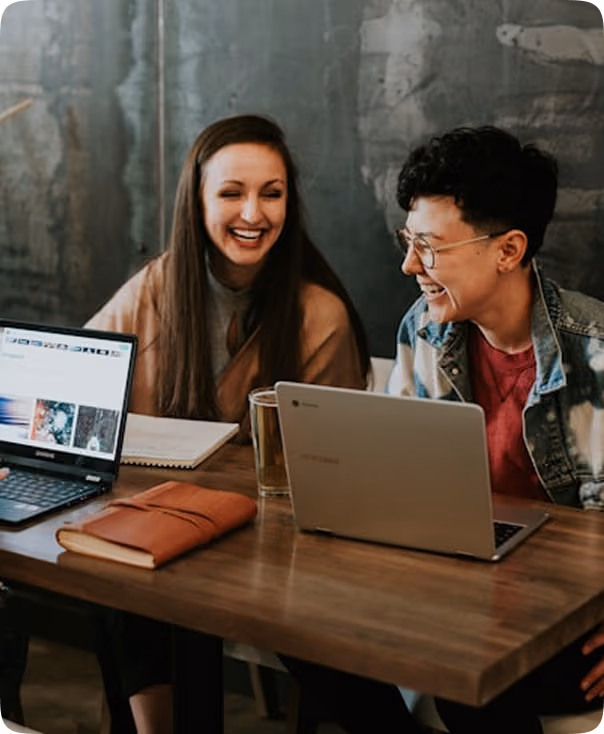 Deux personnes souriantes assises à une table avec des ordinateurs portables et des cahiers lors d'une réunion conviviale.