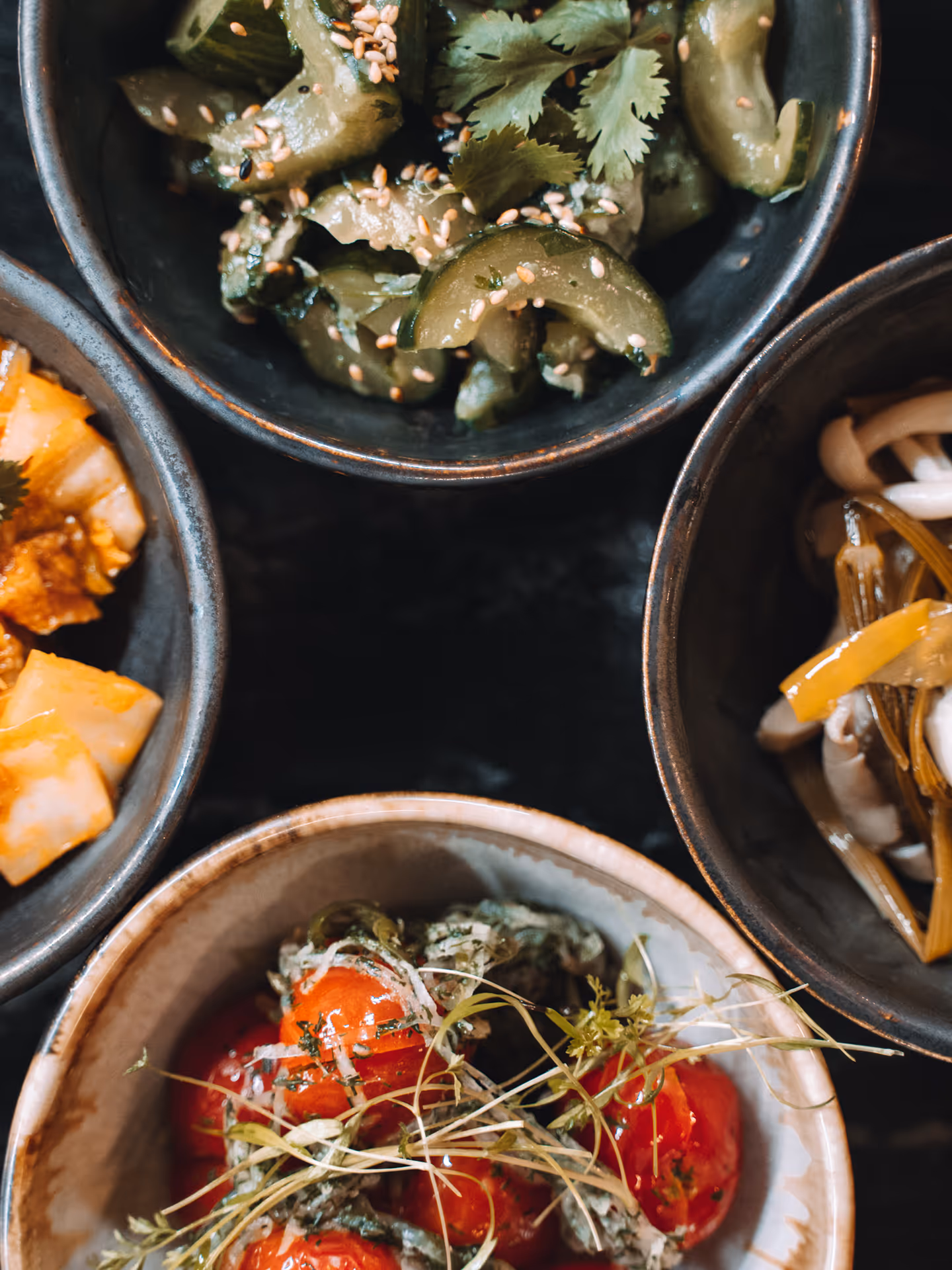 Four bowls of assorted Asian side dishes including pickled cucumbers with sesame seeds, kimchi, marinated vegetables with yellow strips, and cherry tomatoes with herbs.