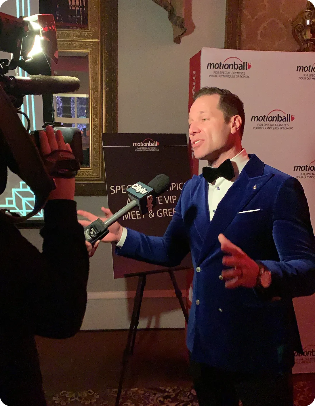Man in a blue velvet tuxedo jacket and bow tie speaking into a CITYNEWS microphone during a Motionball event.