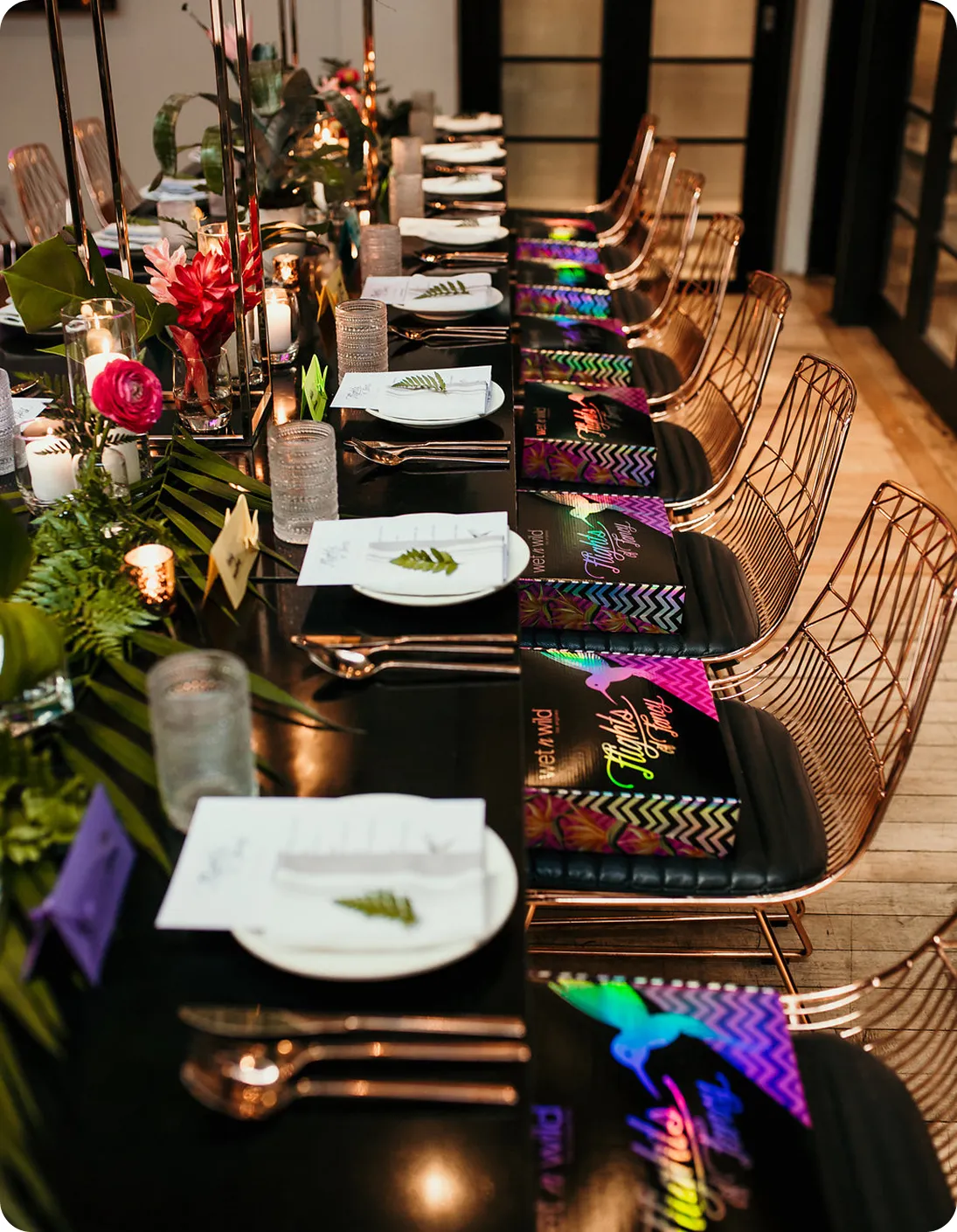 Long dining table set with white plates, silver cutlery, floral decorations, candles, and colorful gift boxes on black cushioned wire chairs.