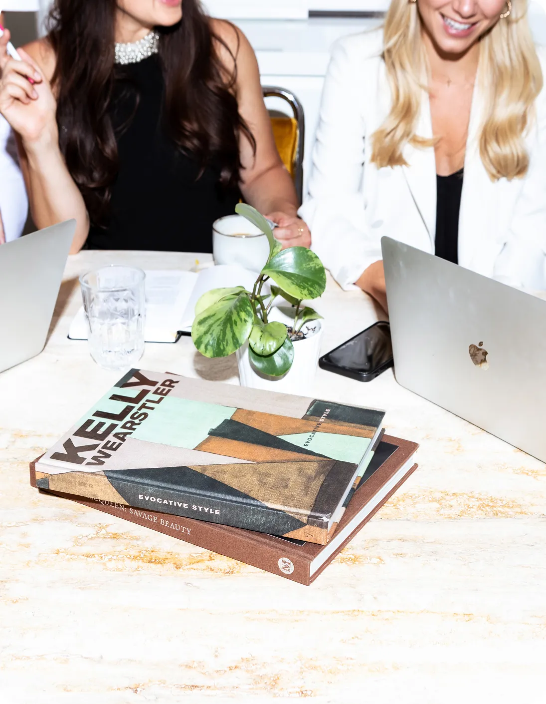 Two women sitting at a table with laptops, a glass cup, a small potted plant, and two stacked books titled 'Kelly Wearstler Evocative Style' and 'Alexander McQueen Savage Beauty'.