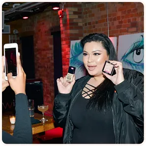 Woman holding two makeup products while another person takes a photo with a smartphone in a dimly lit indoor setting.