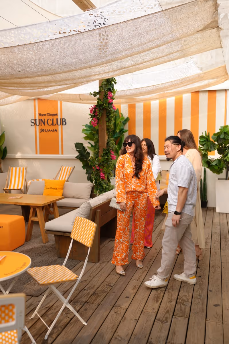 A group of four people socializing in a stylish sunlit lounge area with wooden flooring, orange and white decor, and greenery.