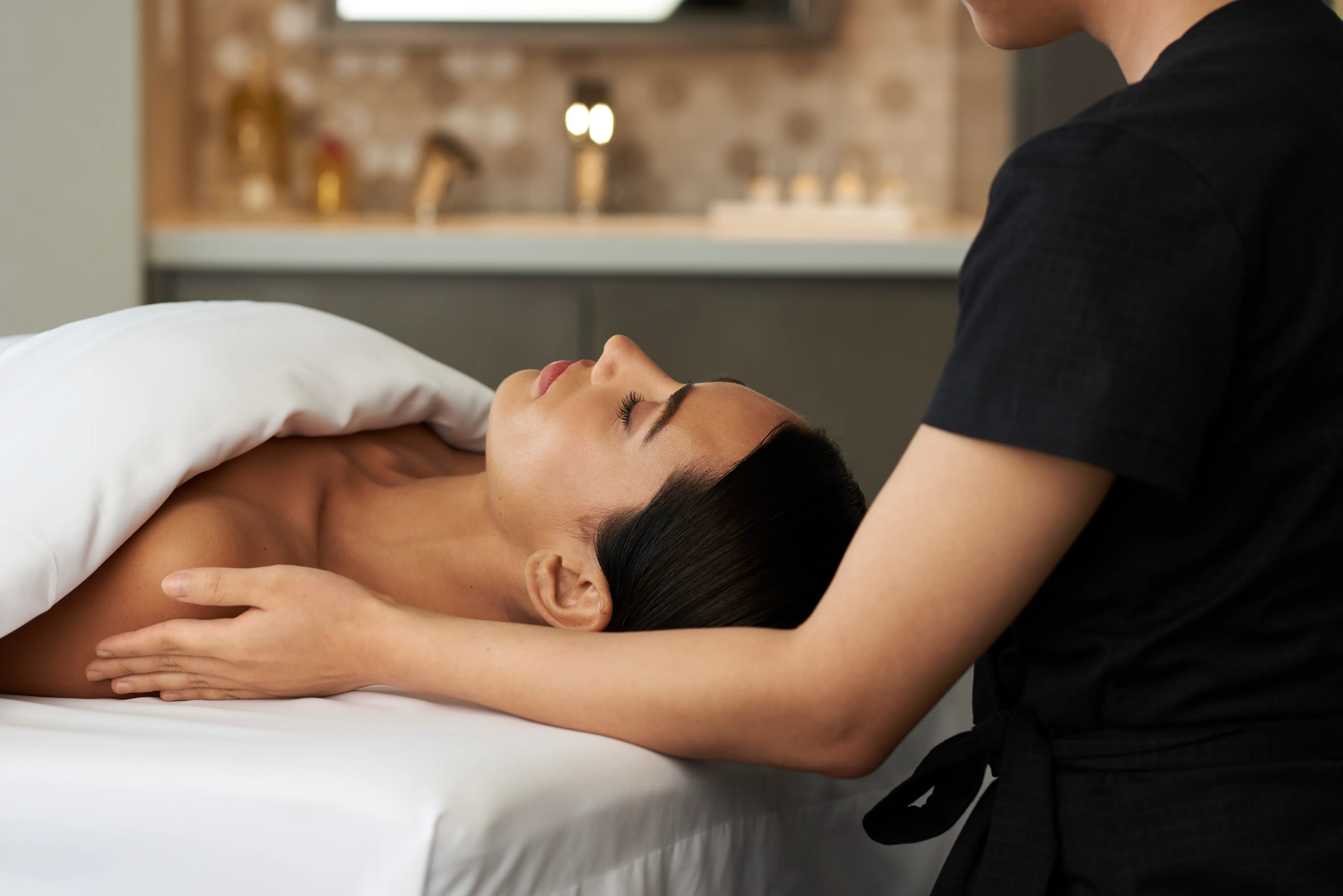 Woman lying on spa bed covered with white towel receiving a facial massage from therapist.
