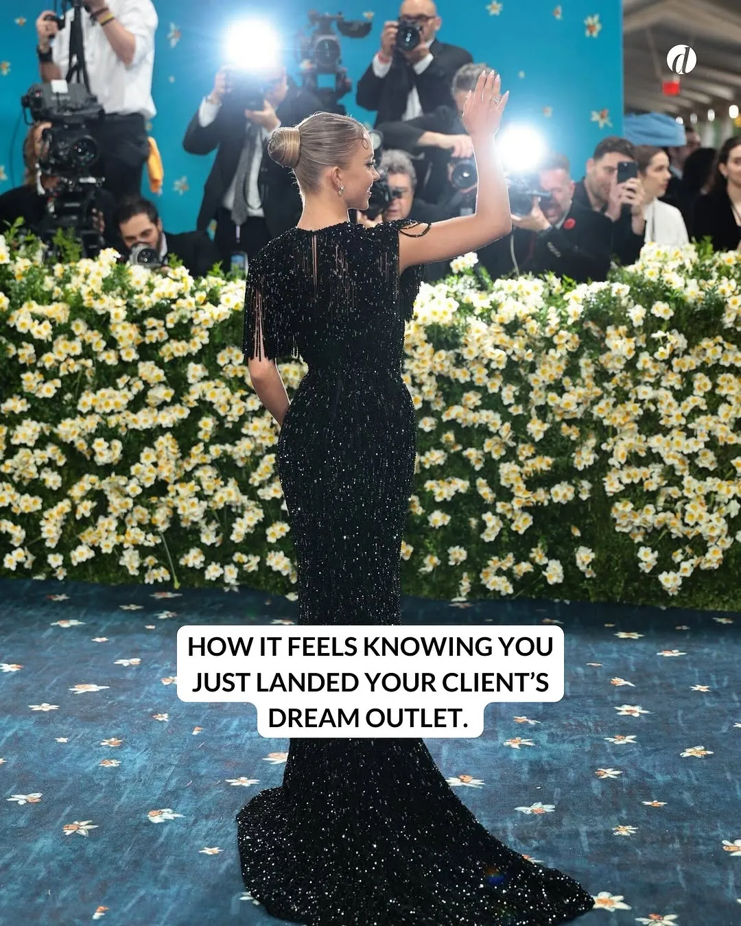 Woman in a sparkling black gown with hair in a bun waves to photographers behind a floral barrier at an event.