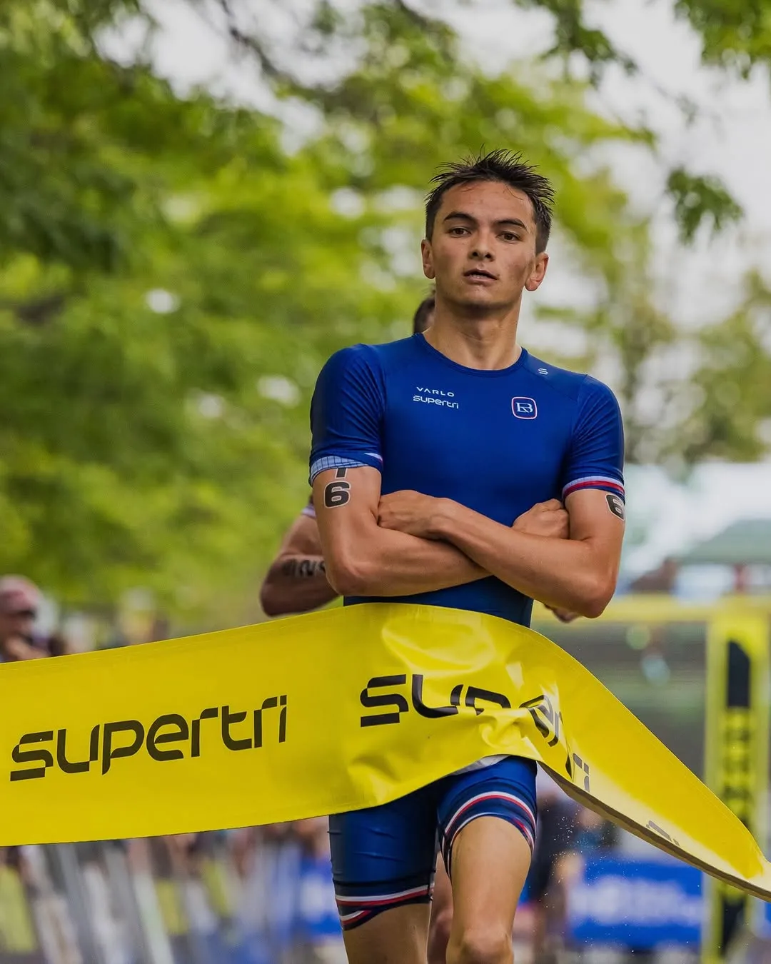 Triathlete crossing the finish line wearing a blue suit with arms crossed and number 6 on both arms.