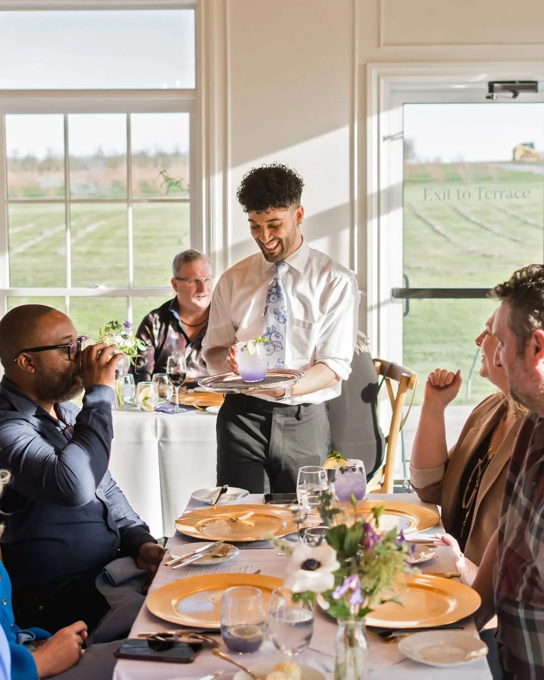 Smiling waiter serving a purple cocktail to a group of people seated at a dining table with gold plates and floral centerpiece.