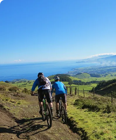 Two mountain bikers riding on a dirt trail overlooking a green landscape and the ocean under a clear blue sky.