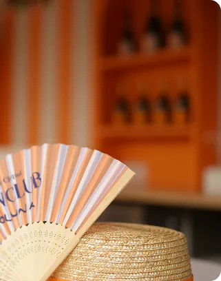 Striped handheld fan resting on a woven straw hat with blurred orange shelves in the background.