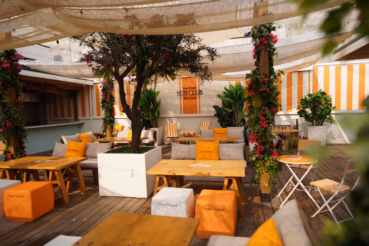 Outdoor lounge at Veuve Clicquot Sun Club Solana with wooden tables, cushioned seating, orange and white decor, and plants under light fabric shades.
