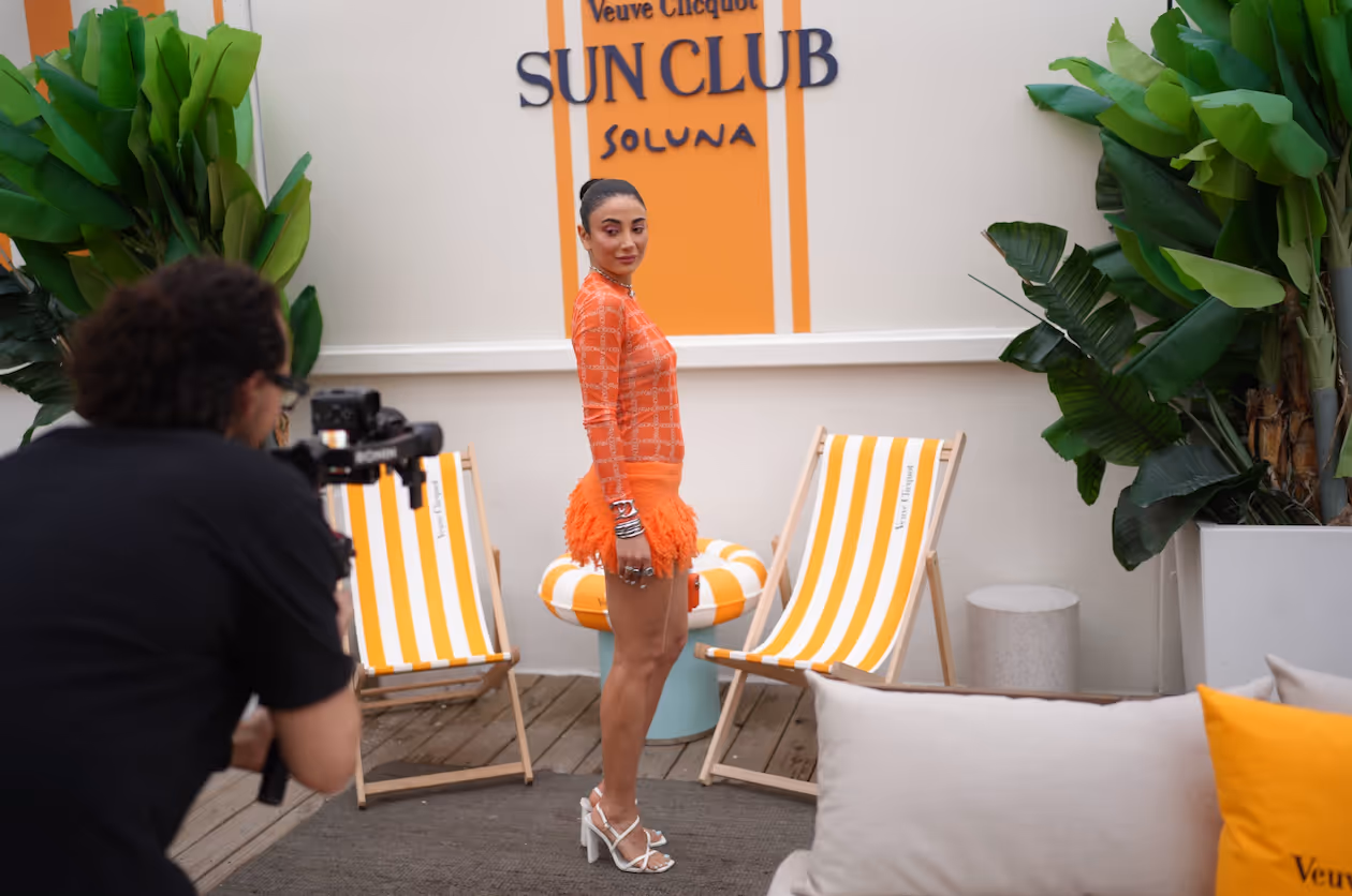 Woman in orange patterned dress and high heels posing for a photographer at Veuve Clicquot Sun Club with striped deck chairs and greenery in the background.