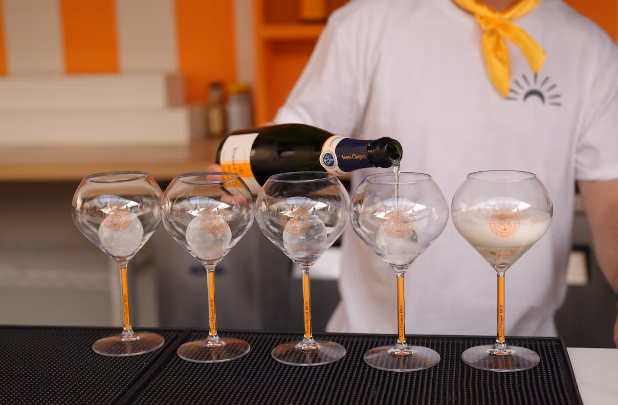 A person pours champagne from a Veuve Clicquot bottle into one of five wine glasses lined up on a bar mat.