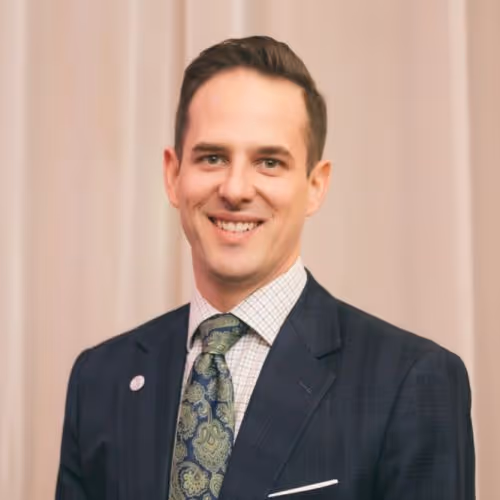 Smiling man wearing a dark suit jacket, white checkered shirt, and patterned tie, standing against a beige curtain background.