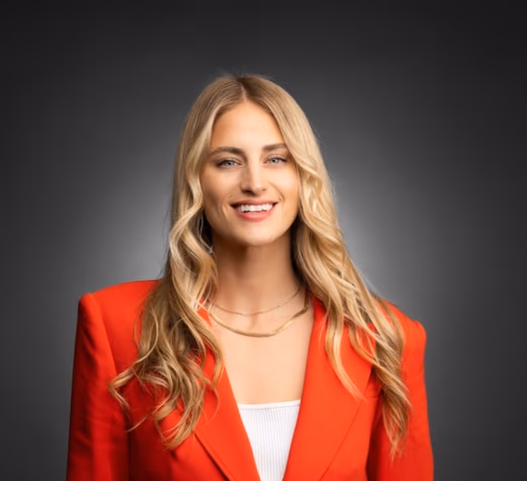 Smiling woman with long blonde hair wearing a red blazer and white top against a dark background.