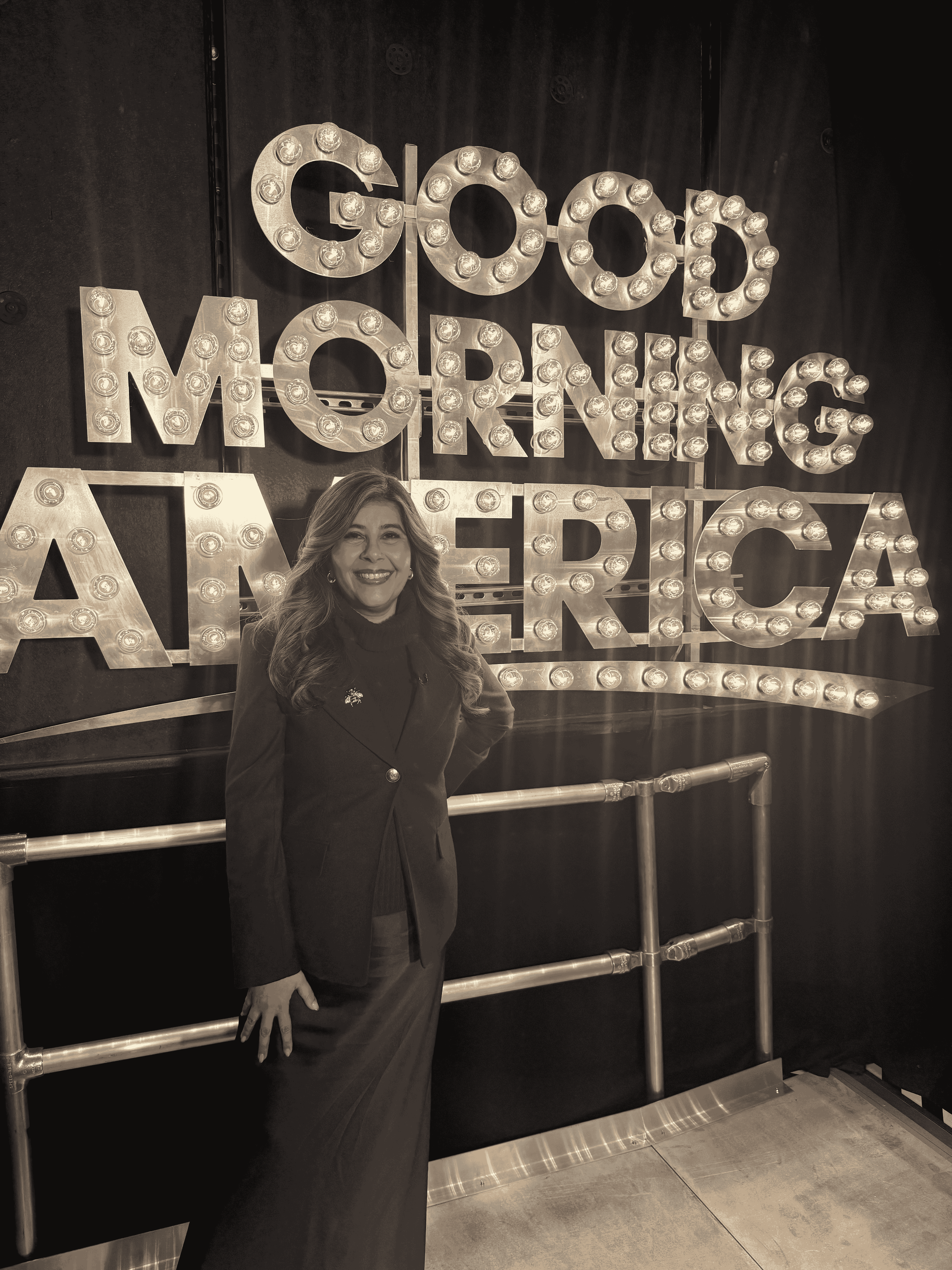 Smiling woman with long hair dressed in a dark blazer and skirt standing in front of a lit Good Morning America sign.