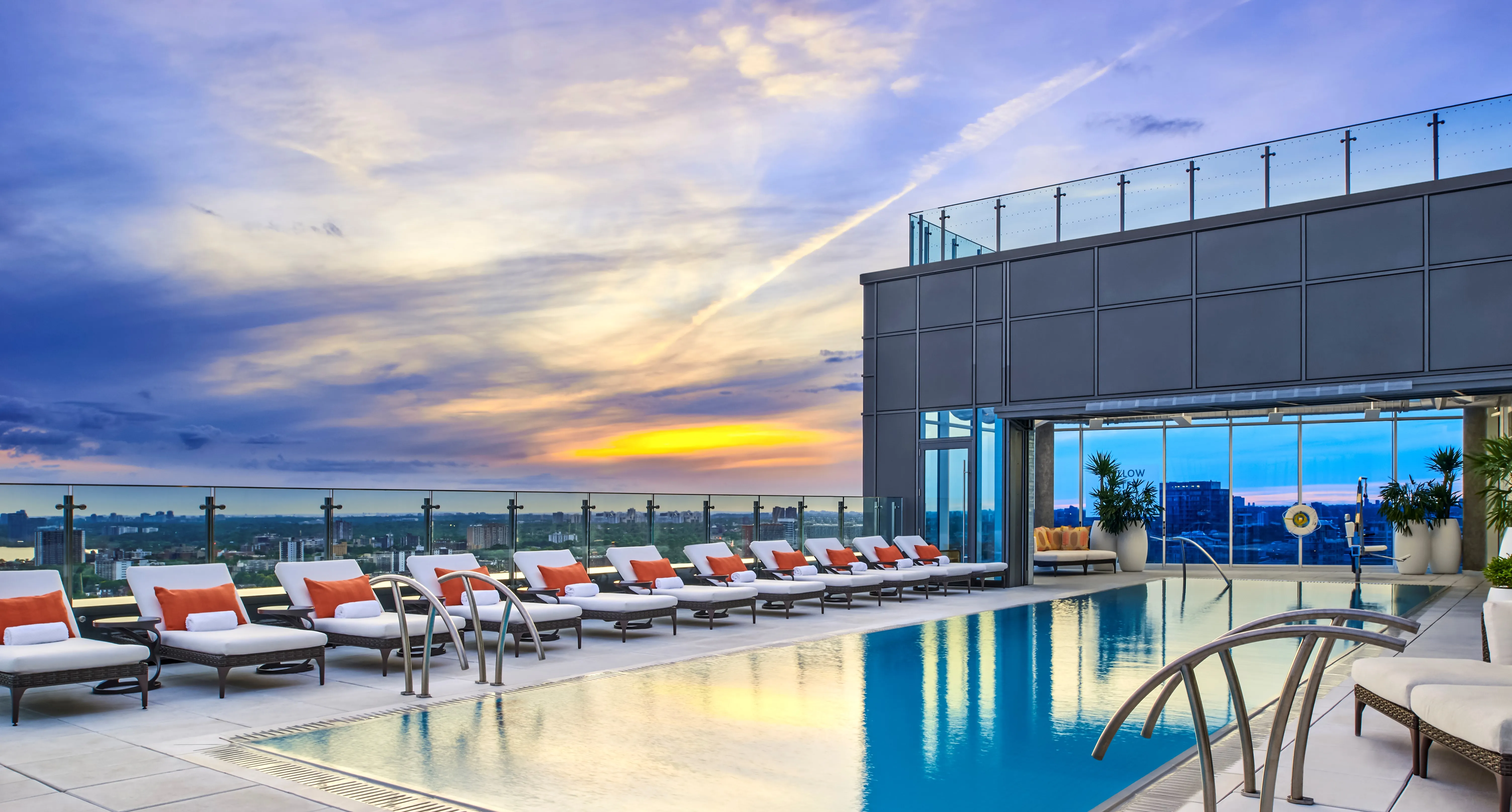 Heated rooftop pool at sunset with cushioned lounge chairs lined up beside it and cityscape in the background.