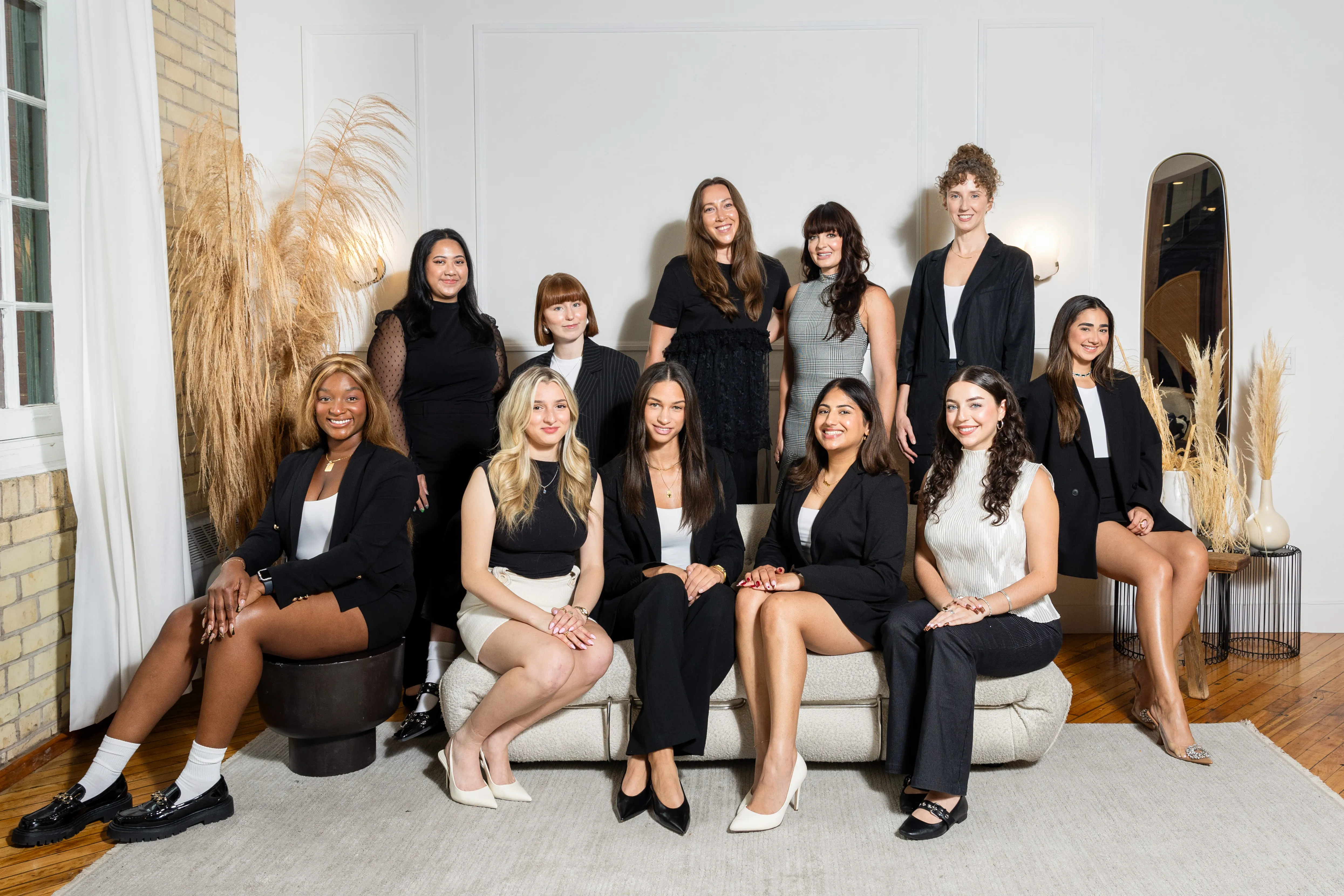 Group of eleven diverse young women dressed in business casual attire posing in a bright, modern room with neutral decor and pampas grass accents.