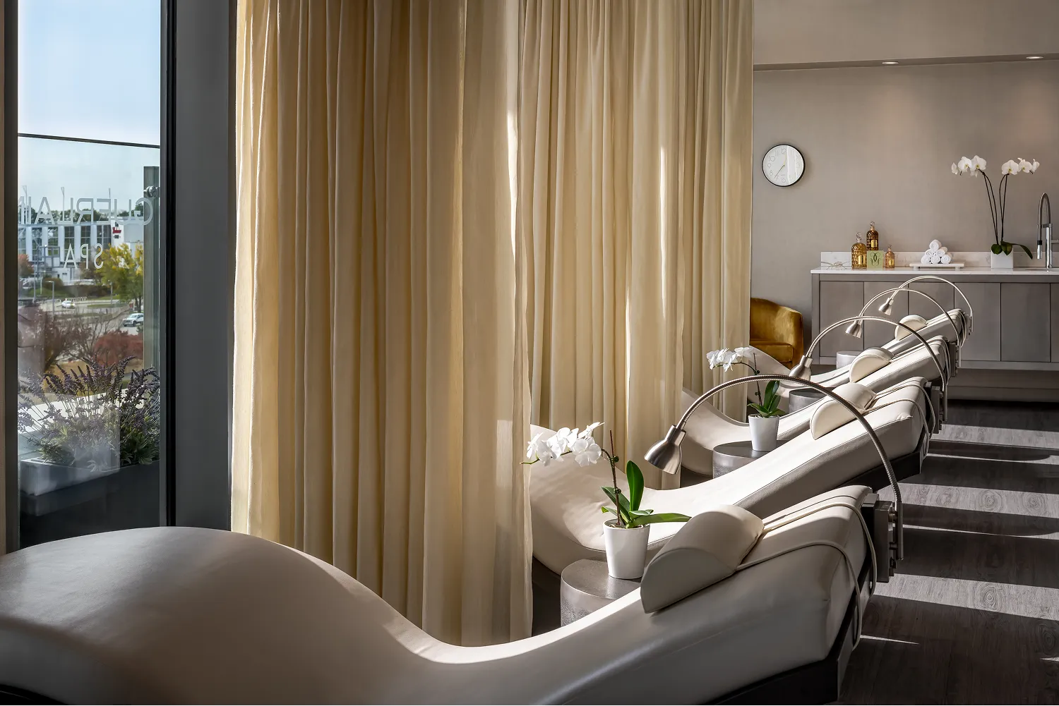 Relaxation area in a spa with reclining white chairs, small tables holding white orchid plants, beige curtains, and a counter with towels and spa products.