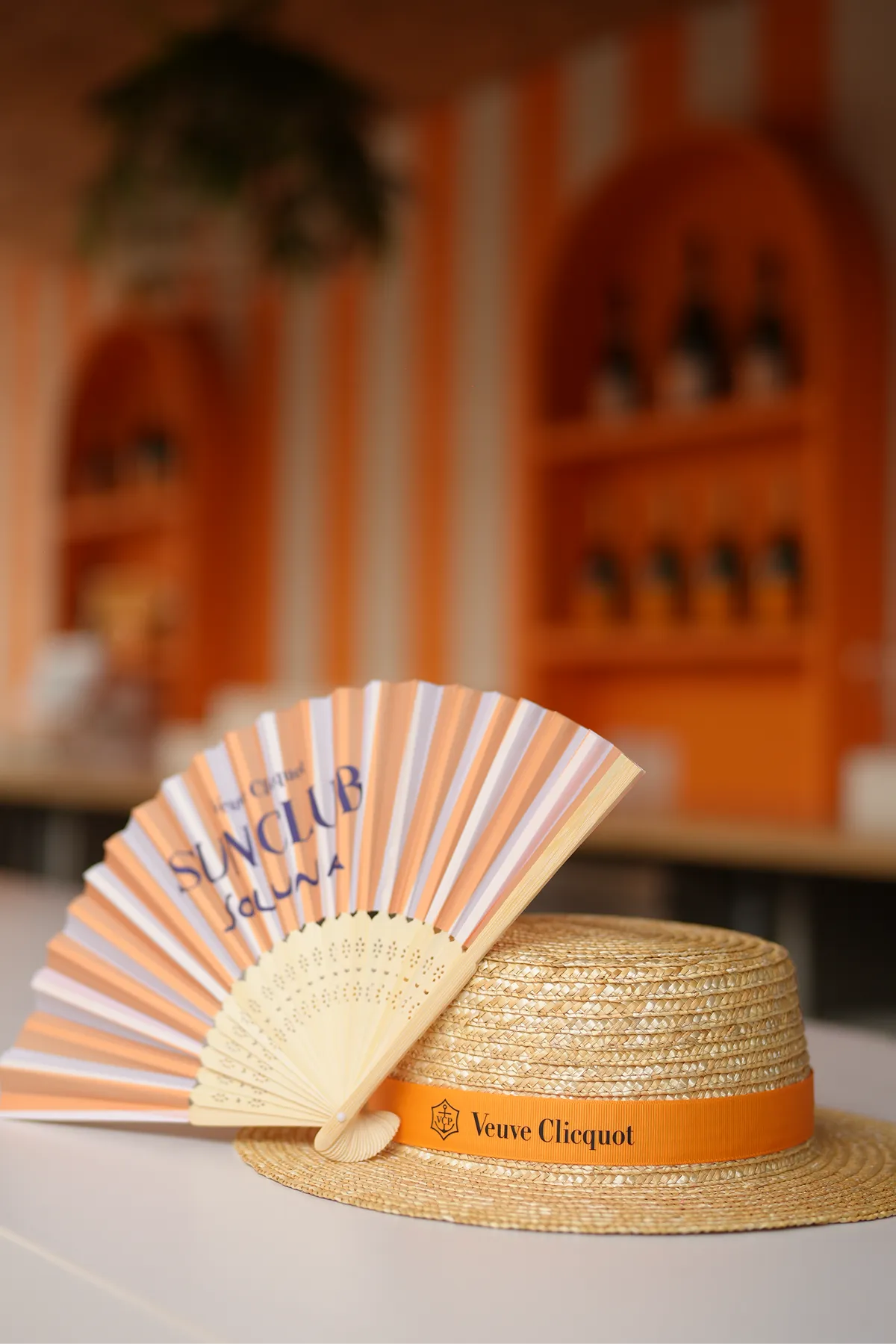 Straw boater hat with an orange Veuve Clicquot band and a pink and white striped hand fan with Sunclub Soluna text resting on a table.