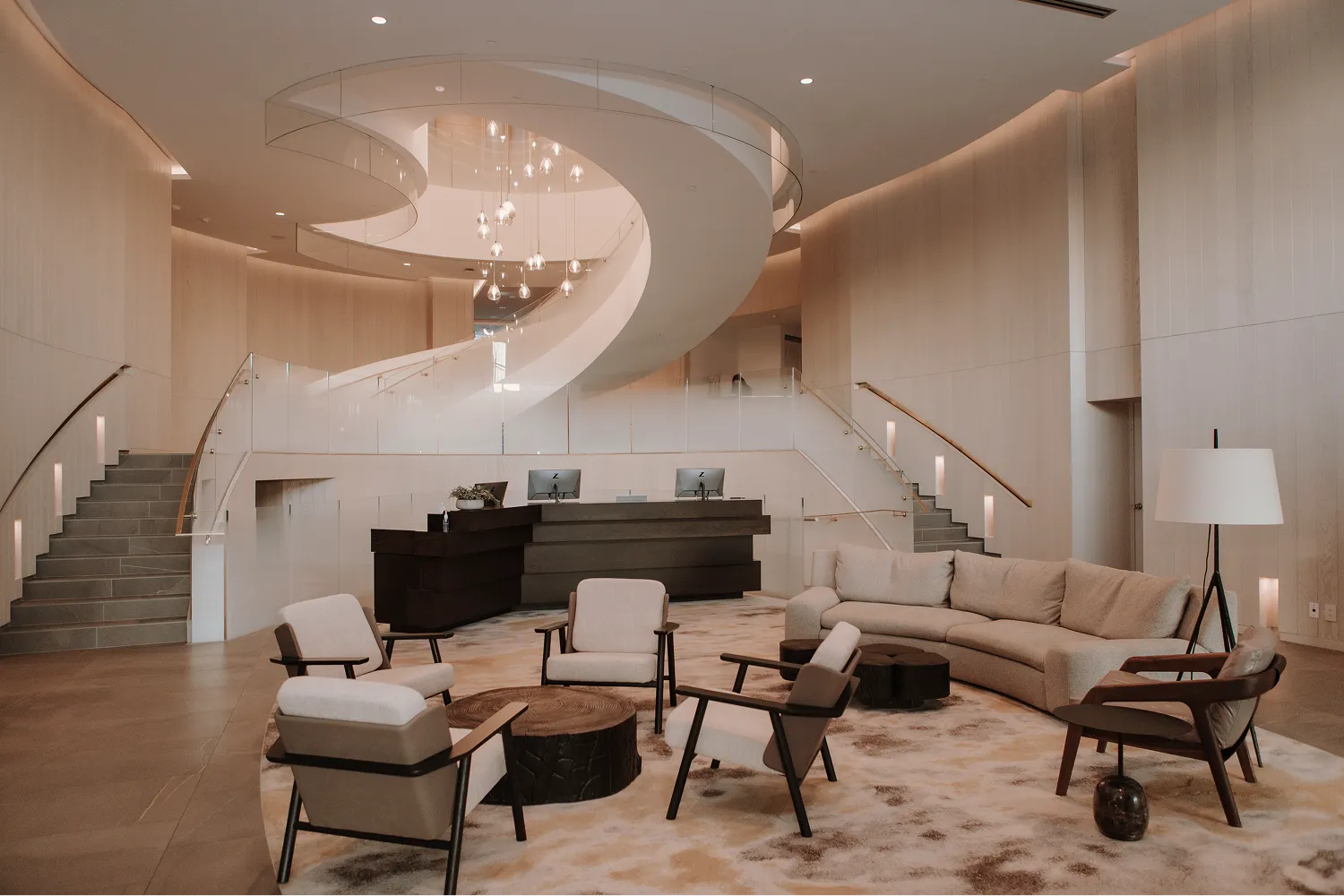 Modern hotel lobby with curved white staircase, beige sofas, armchairs, and a wooden reception desk.