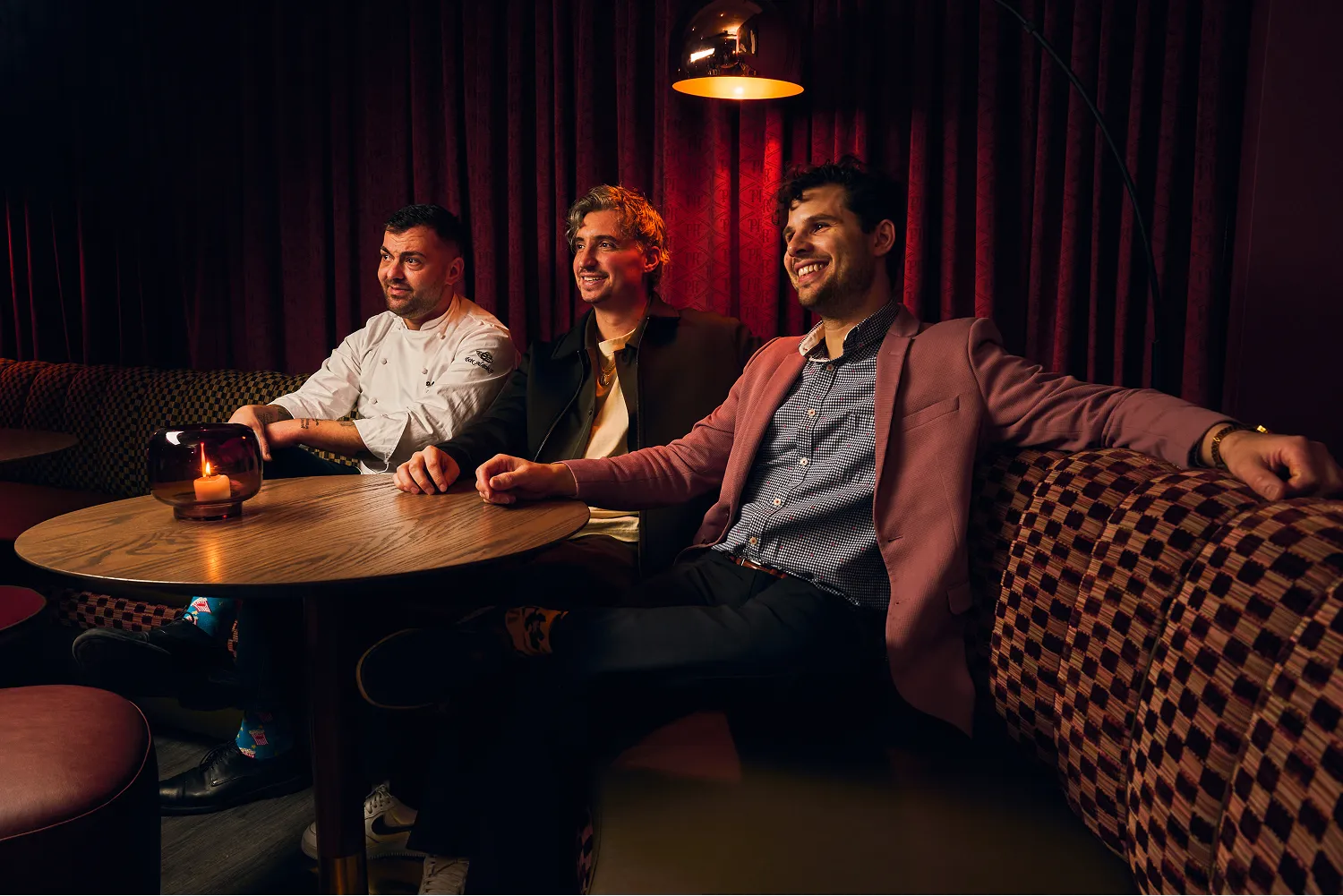 Three men sitting in a cozy dimly lit lounge with red curtains, smiling and relaxing around a wooden table with a lit candle.