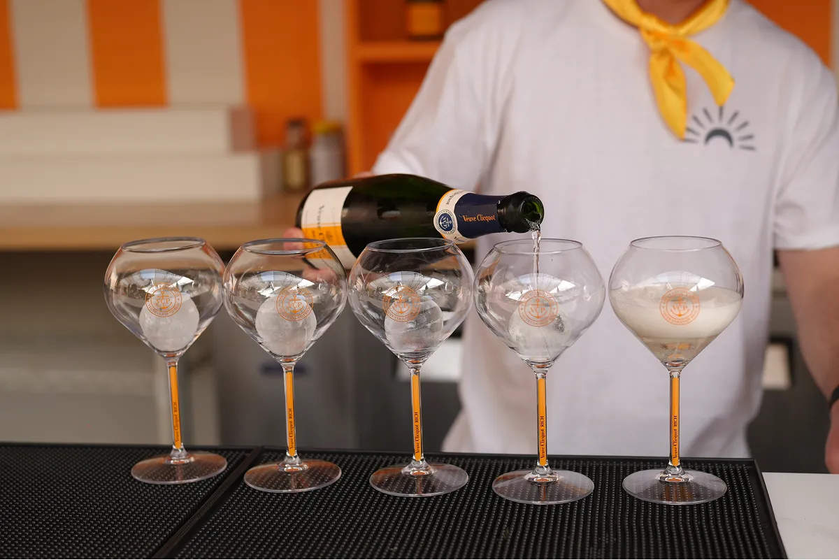 Person pouring Veuve Clicquot champagne into one of five glasses each containing an ice ball on a black bar mat.