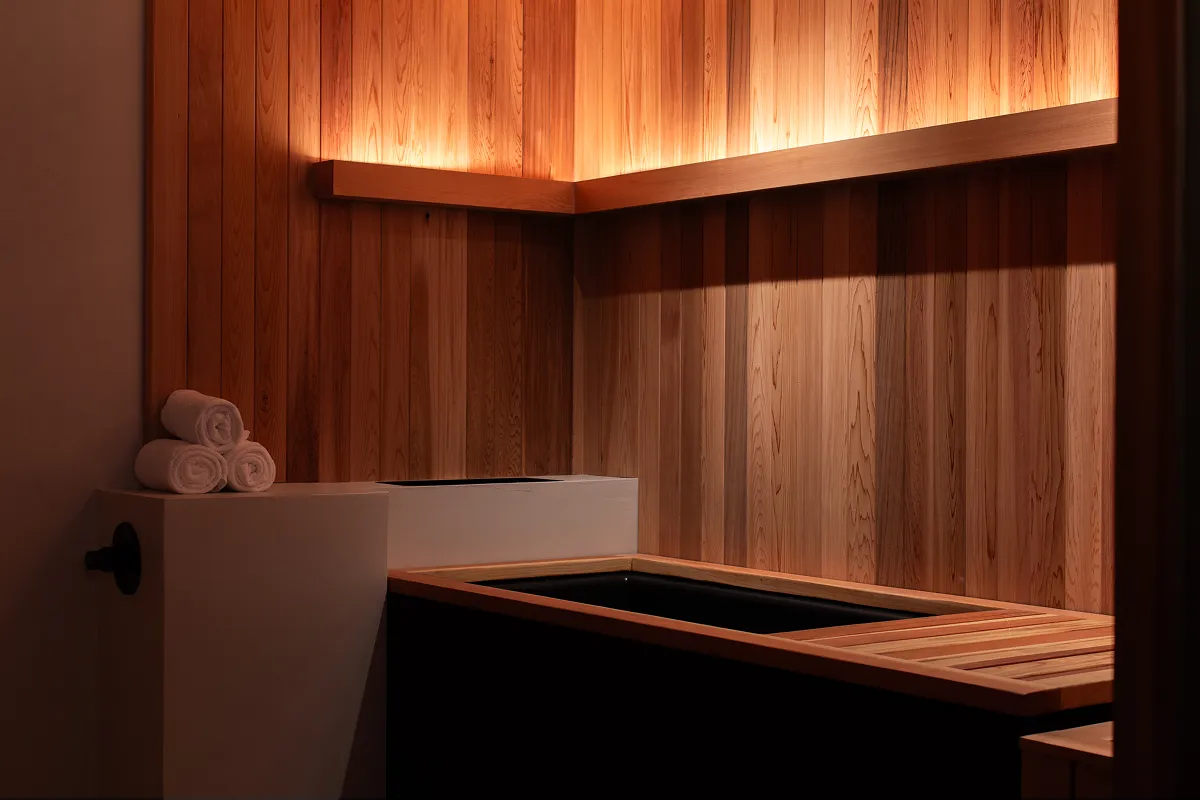 Modern wooden soaking tub area with ambient lighting and rolled white towels on a small countertop.